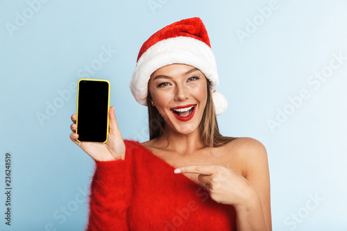 Young woman in christmas hat posing isolated over blue wall background using mobile phone showing empty display.