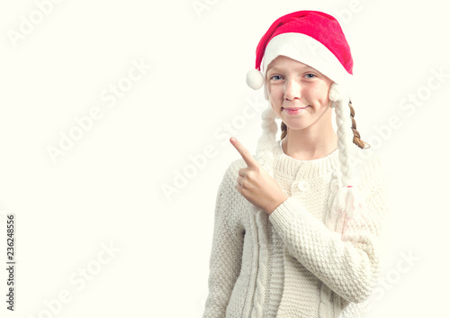 Portrait of a teenager girl in a red Santa Claus hat pointing with his finger towards the left while standing on a light background. Place for text. photo