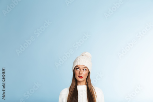Thinking young woman wearing winter hat posing isolated over blue wall background.