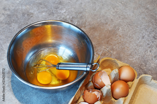 Metal bowl with three raw eggs for preparing omelet for breakfast and cardboard package with empty cracked egg shells and two brown chicken eggs on grunge grey kitchen table background.