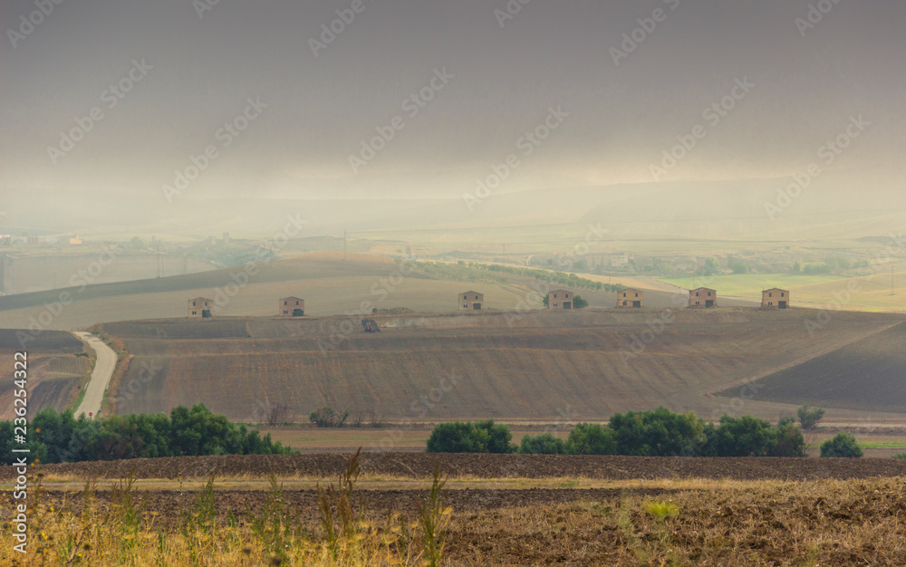 Between Apulia and Basilicata: hilly rural landscape with farmhouses on plowed land dominated by clouds, Italy. Country road through plowed field.