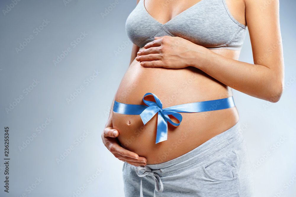 Close up of pregnant woman with blue ribbon touching her belly on grey background. Pregnancy, maternity, preparation and expectation concept Close up of pregnant woman with blue ribbon touching her belly on grey background. Pregnancy, maternity, preparation and expectation concept