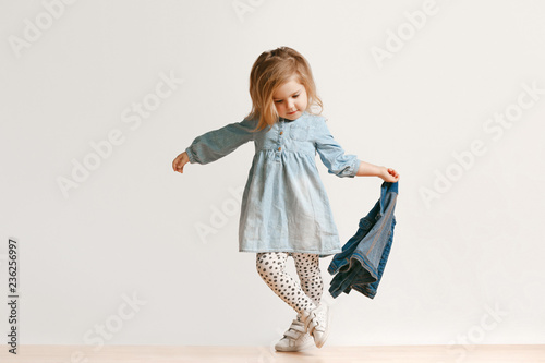 Full length portrait of cute little kid girl in stylish jeans clothes looking at camera and smiling, standing against white studio wall. Kids fashion concept
