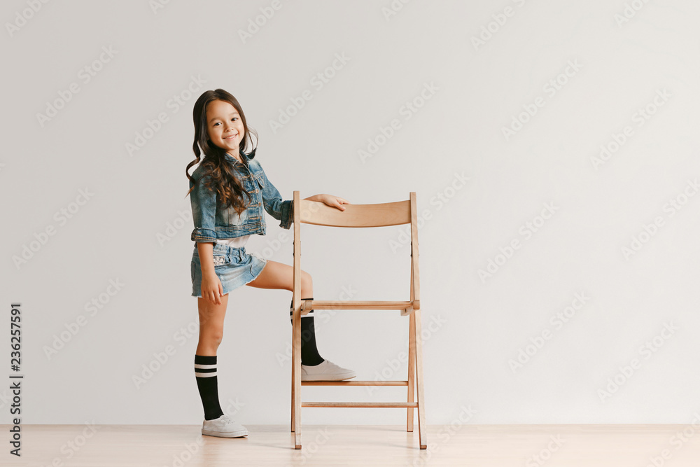 Full length portrait of cute little kid girl in stylish jeans clothes looking at camera and smiling, standing against white studio wall. Kids fashion concept Full length portrait of cute little kid girl in stylish jeans clothes looking at camera and smiling, standing against white studio wall. Kids fashion concept