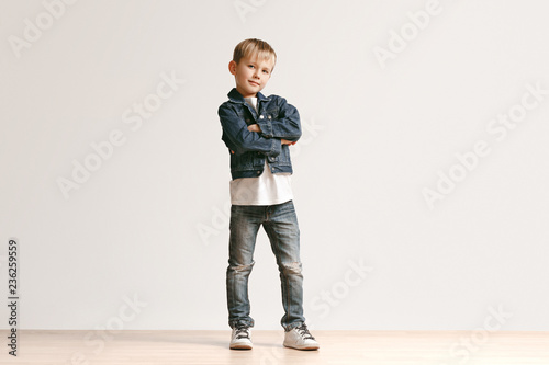 The portrait of cute little kid boy in stylish jeans clothes looking at camera against white studio wall. Kids fashion concept photo