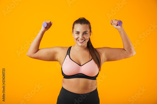 Portrait of beautiful big sportswoman in sportive bra lifting dumbbells, isolated over yellow background