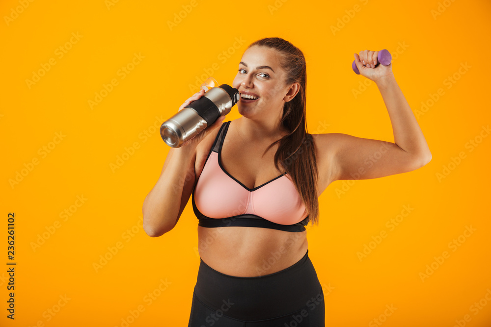 © Drobot Dean - Portrait of young chubby woman in sportive bra drinking water from thermos while lifting dumbbell, isolated over yellow background © Drobot Dean - Portrait of young chubby woman in sportive bra drinking water from thermos while lifting dumbbell, isolated over yellow background