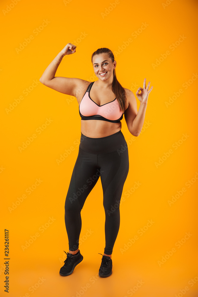Full length portrait of beautiful chubby woman in sportive bra showing her bicep, isolated over yellow background - obrazy, fototapety, plakaty 