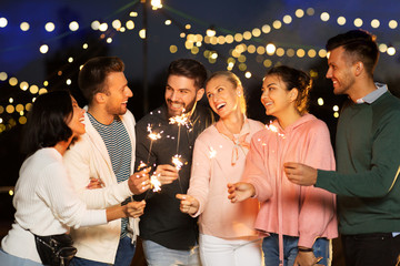 leisure, celebration and people concept - happy friends with sparklers at rooftop party at night with blurred bokeh lights