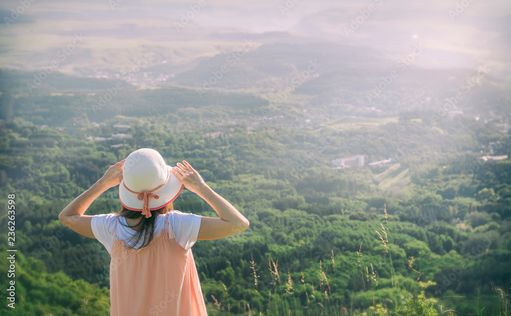 Girl with a a hat standing with her back against a background of green mountains.
