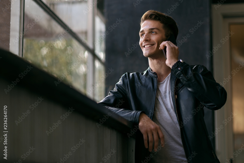 Happy man standing by the window and talking to someone over smart phone