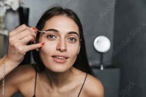 Photo of european woman 20s with long dark hair standing in bathroom, and plucking eyebrows with tweezers