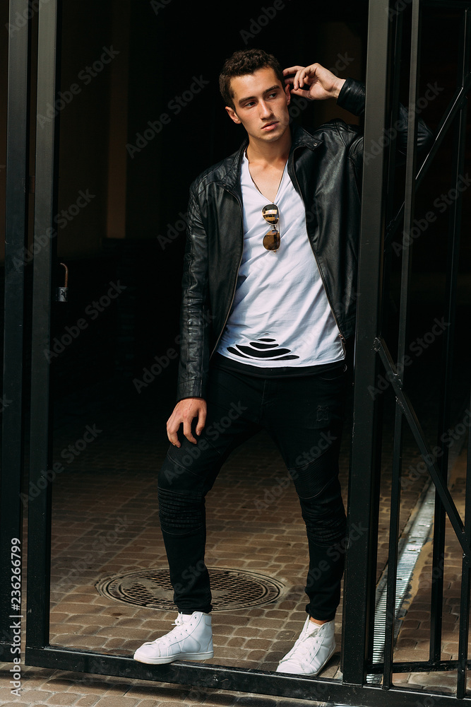 young, handsome guy in a white T-shirt, black, leather jacket and black  pants is on the street, on the T-shirt sunglasses. model, city walk, live  style. in white sneakers Stock Photo