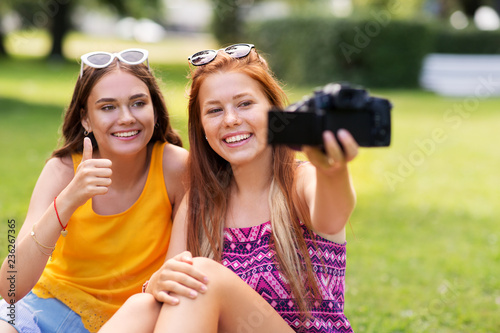blogging, technology and leisure concept - teenage girls, friends or bloggers recording video by camera in summer park and showing thumbs up