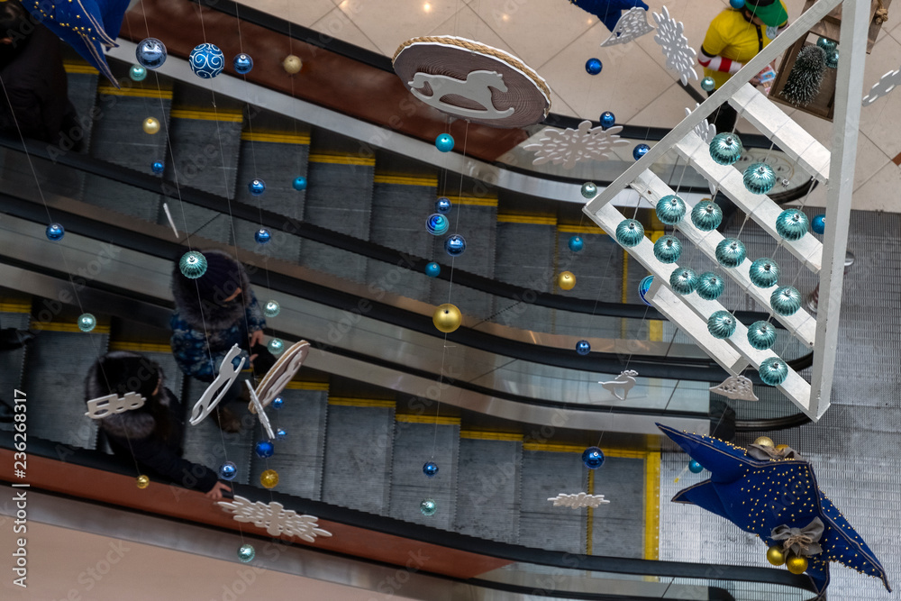 Decorating a shopping center or airport in the Christmas style. Balls,  lanterns, a horse and other toys hang on thin threads above the escalator.  Concept for the New Year. Stock Photo