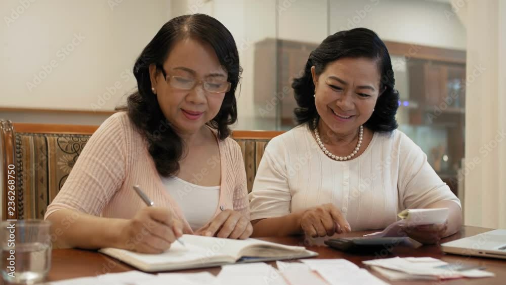 Elegant elderly Asian women sitting at table with papers and receipts, counting sales on calculator, making notes in register and smiling with satisfaction Elegant elderly Asian women sitting at table with papers and receipts, counting sales on calculator, making notes in register and smiling with satisfaction