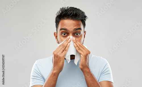 health problem, rhinitis and allergy concept - unhealthy indian man with paper napkin blowing nose over grey background
