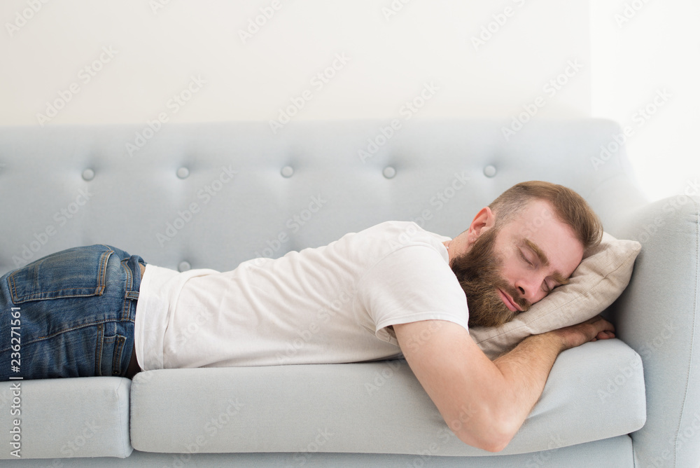 Handsome bearded man lying and sleeping on stomach on sofa. Young guy dozing. Rest concept.