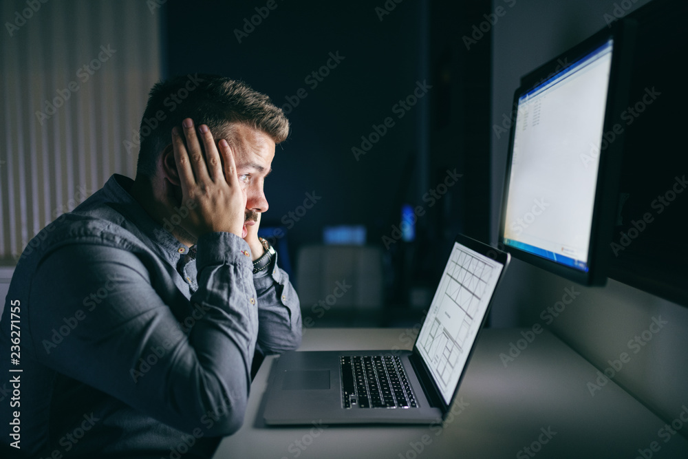 Tired young Caucasian employee holding head in hands and looking at computer monitor while sitting late at night in the office. Tired young Caucasian employee holding head in hands and looking at computer monitor while sitting late at night in the office.