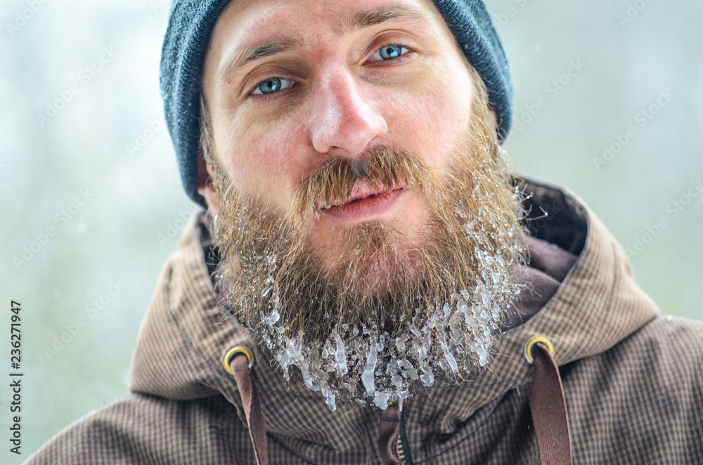 Portrait of a looking at camera young man in a gray hat and a brown jacket with a long beard covered with icicles. Men winter joys concept Portrait of a looking at camera young man in a gray hat and a brown jacket with a long beard covered with icicles. Men winter joys concept