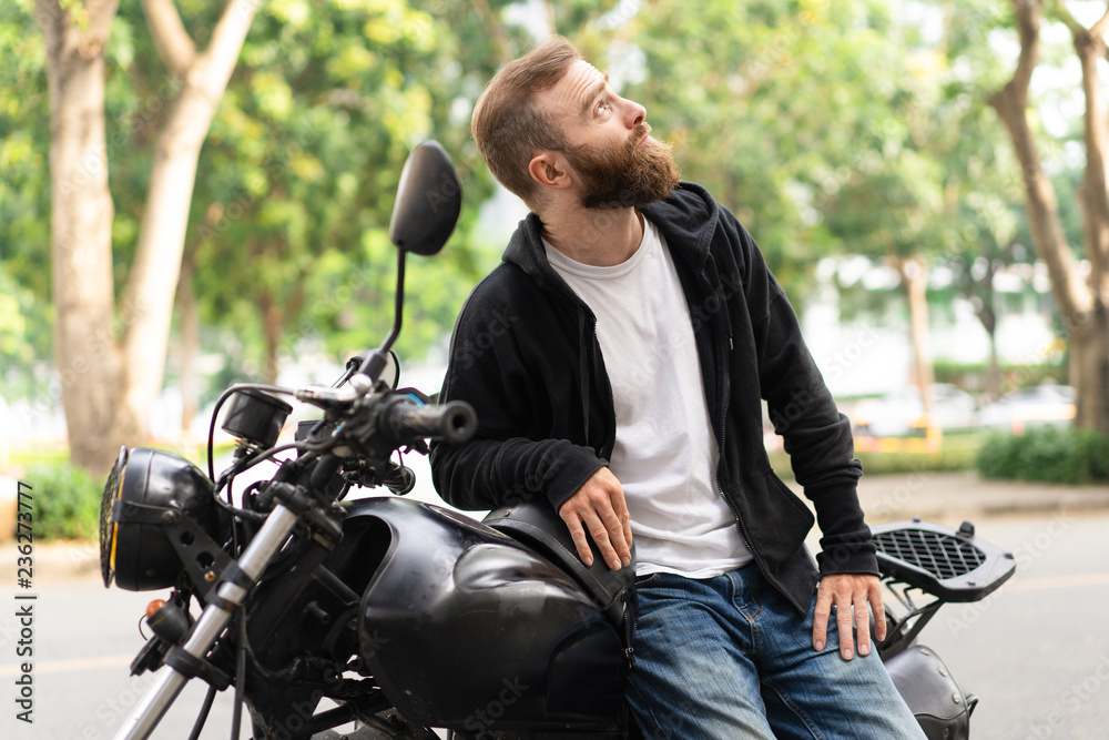 Portrait of confident biker resting in his motorbike looking up. Young Caucasian man sitting on motorcycle outdoors. Biker culture concept