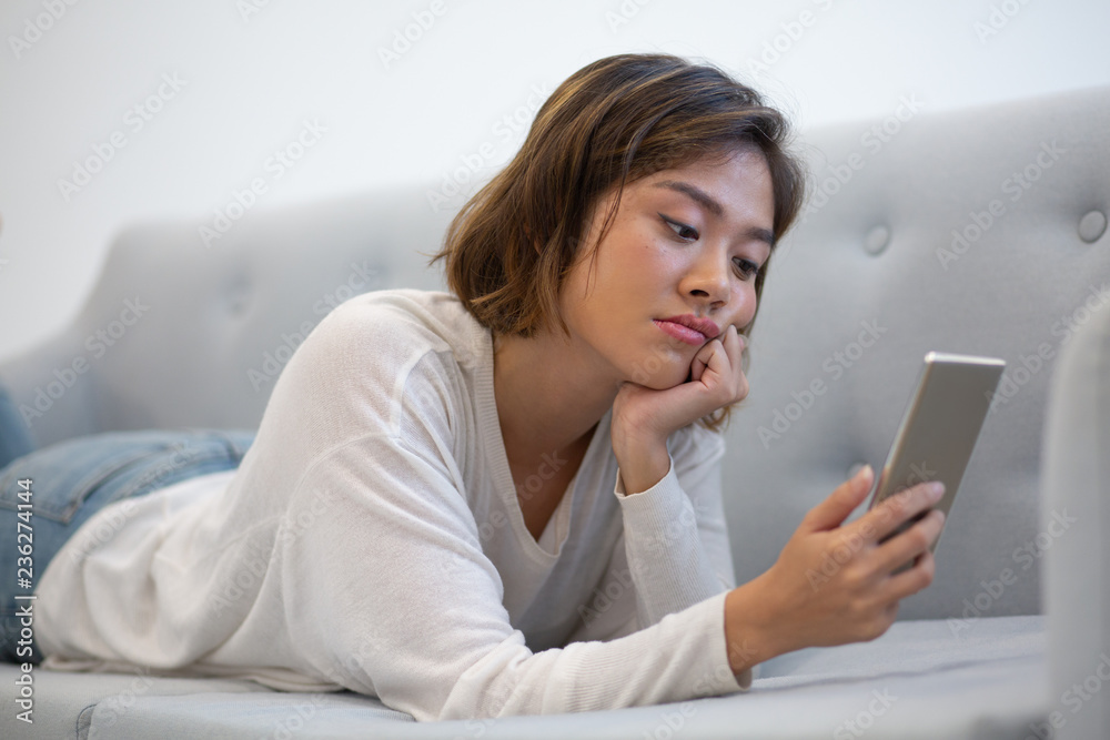 Bored Asian phone user browsing internet. Young woman in casual lying on couch, leaning cheek on hand and using smartphone. Boredom concept Bored Asian phone user browsing internet. Young woman in casual lying on couch, leaning cheek on hand and using smartphone. Boredom concept