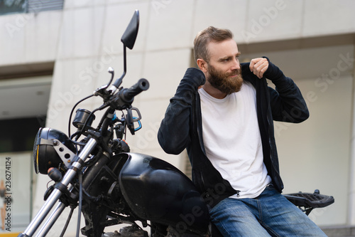 Smiling young biker with motorcycle putting on hoodie. Portrait of Caucasian bearded man sitting on motorbike outdoors. Biker culture concept