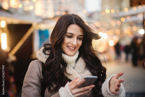 Close up of young smiling beautiful Caucasian woman using smart phone on the street on cold weather.