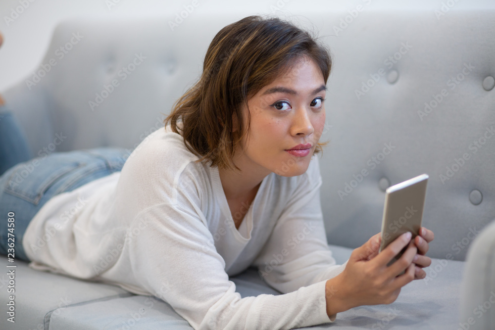 Cute pensive Asian girl browsing internet. Young woman in casual lying on couch, using smartphone and looking at camera. Phone addiction concept Cute pensive Asian girl browsing internet. Young woman in casual lying on couch, using smartphone and looking at camera. Phone addiction concept