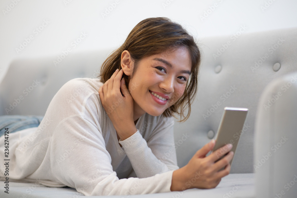 Happy Asian phone user satisfied with new mobile app. Young woman in casual resting on couch, holding smartphone and smiling at camera. Mobile app concept Happy Asian phone user satisfied with new mobile app. Young woman in casual resting on couch, holding smartphone and smiling at camera. Mobile app concept
