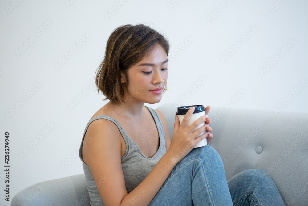 Pensive Asian girl drinking morning coffee. Young woman in casual sleeveless shirt sitting on couch and holding carton cup. Morning concept Pensive Asian girl drinking morning coffee. Young woman in casual sleeveless shirt sitting on couch and holding carton cup. Morning concept