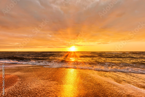 Fototapeta Naklejka Na Ścianę i Meble -  scenic sunset over sea with setting sun rays coming through dramatic clouds on orange blue sky reflecting on water panoramic aerial nature dusk atmosphere evening landscape background