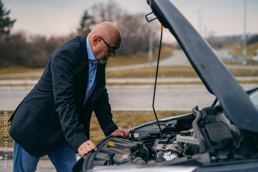 Bearded senior adult man standing in front of opened hood of his car and trying to fix engine. Bearded senior adult man standing in front of opened hood of his car and trying to fix engine.