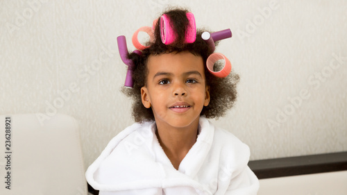 Black girl with curlers with hairpins in her hair. A little girl in a white coat spun pink curlers into her hair.