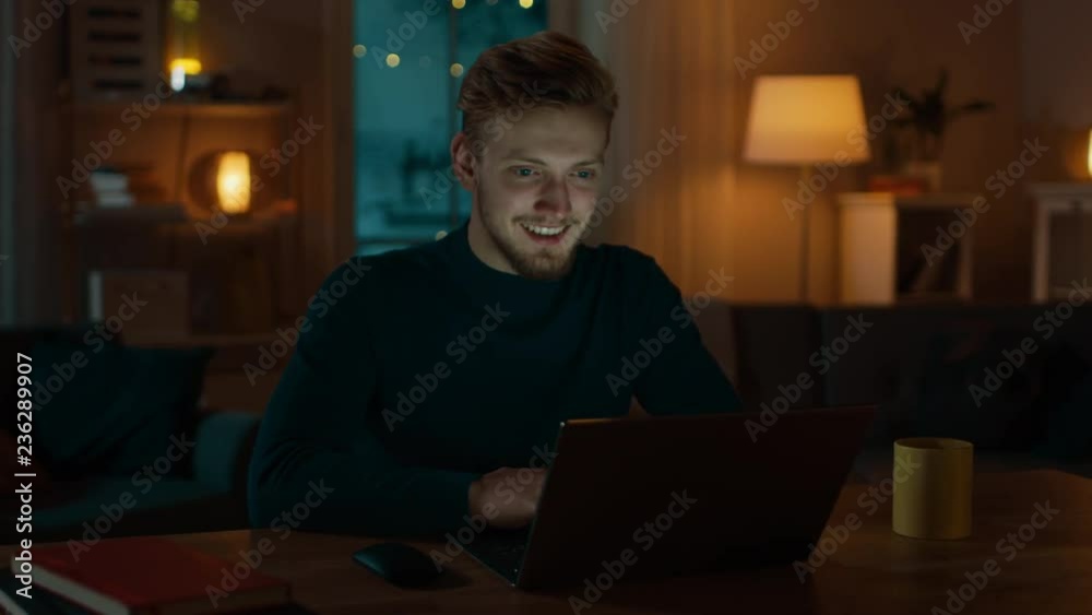 Handsome Smiling Man Works on a Laptop Computer while Sitting at His Desk at Home. Cozy Living Room with Warm Evening Lighting Turned on. Handsome Smiling Man Works on a Laptop Computer while Sitting at His Desk at Home. Cozy Living Room with Warm Evening Lighting Turned on.