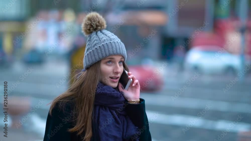 A young girl walking down the street and talking on the phone. Near ride cars. Winter and cold. Beautiful hat. A young girl walking down the street and talking on the phone. Near ride cars. Winter and cold. Beautiful hat.