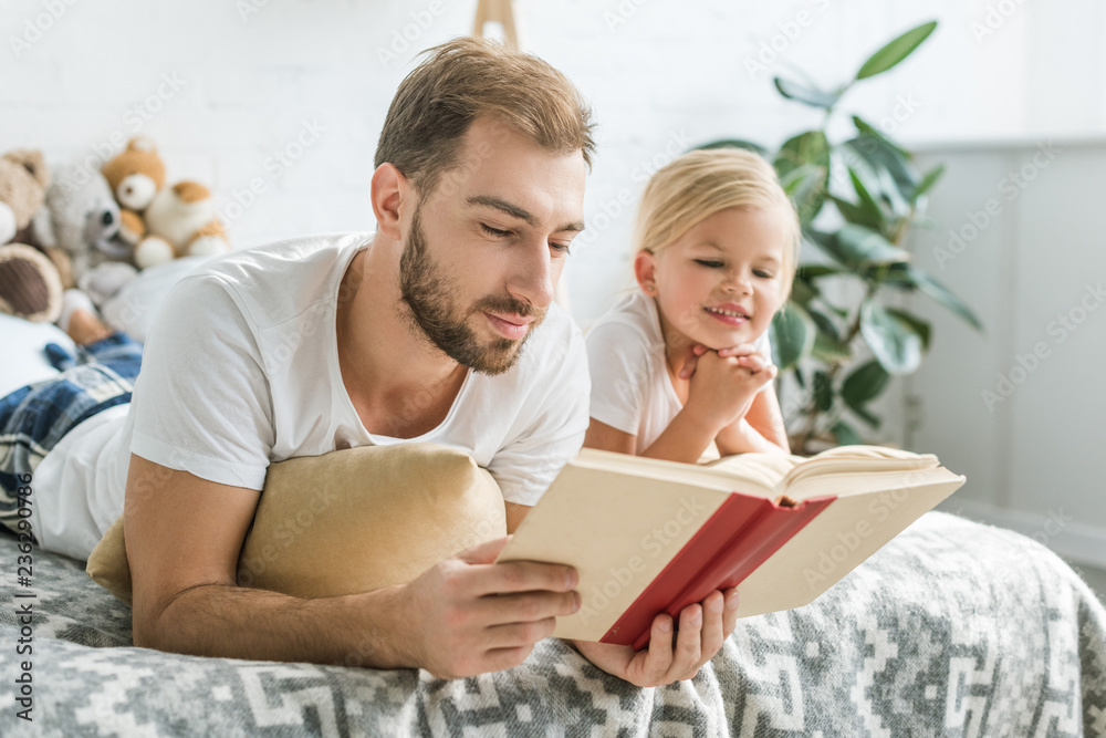 happy father and cute smiling daughter lying on bed and reading book happy father and cute smiling daughter lying on bed and reading book