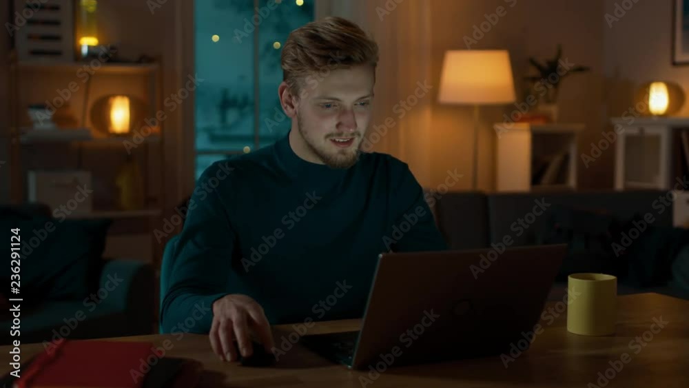 Handsome Smiling Man Works on a Laptop while Sitting at His Desk at Home. Young Freelancer Works on Computer in His Cozy Living Room with Warm Evening Lighting Turned on. Handsome Smiling Man Works on a Laptop while Sitting at His Desk at Home. Young Freelancer Works on Computer in His Cozy Living Room with Warm Evening Lighting Turned on.
