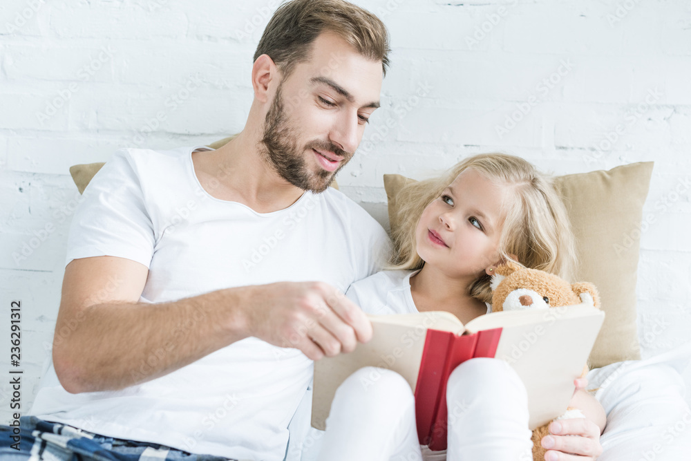 © LIGHTFIELD STUDIOS - father and cute little daughter with teddy bear reading book together on bed