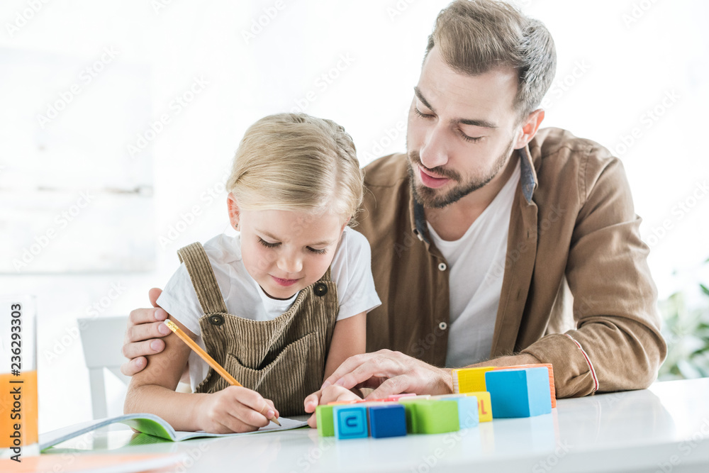father looking at cute little daughter writing in workbook and learning at home father looking at cute little daughter writing in workbook and learning at home