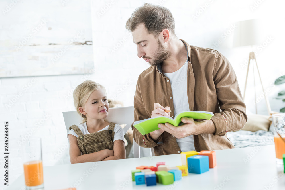 father writing in workbook and looking at little daughter learning math with colorful cubes father writing in workbook and looking at little daughter learning math with colorful cubes