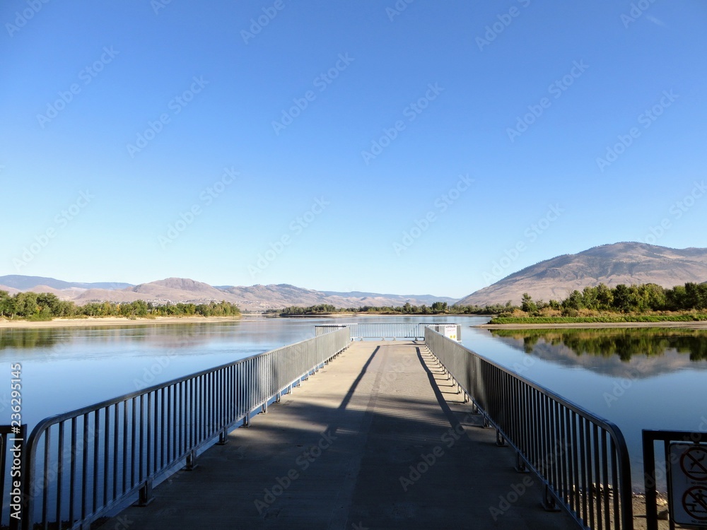 A walking path looking out towards the North Thompson River, in Kamloops, British Columbia, Canada.  Beautiful reflection of the clouds, sky, and mountain in the river.