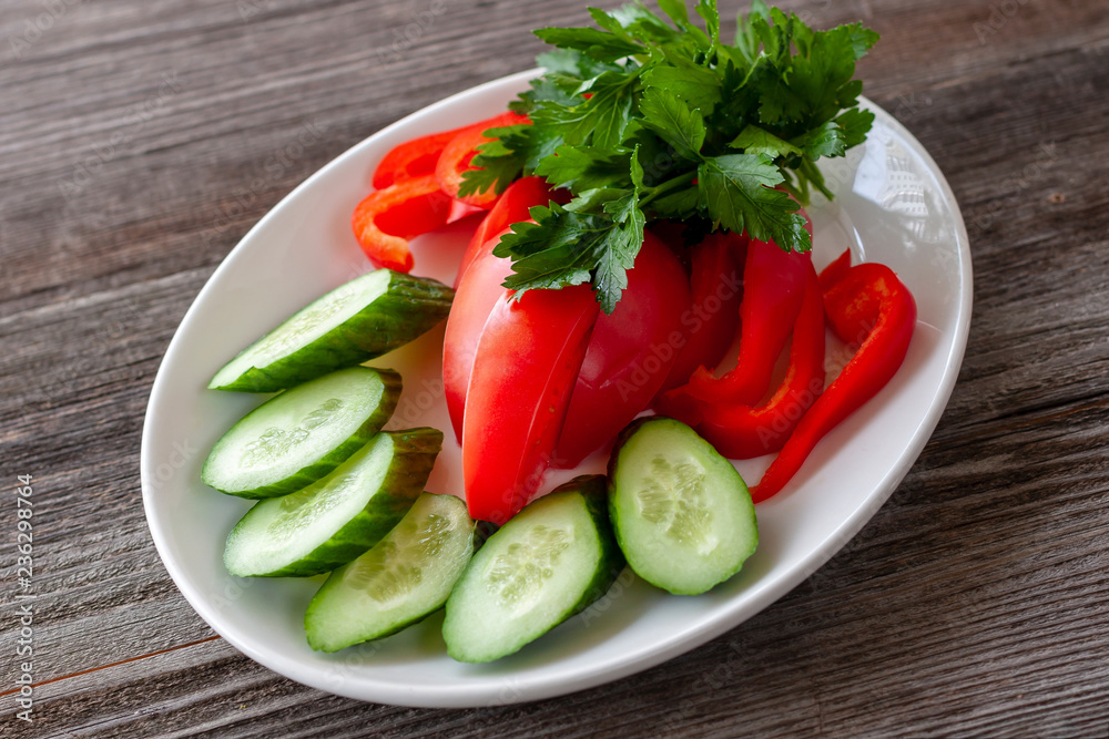 Sliced fresh vegetables, sweet peppers and cucumbers on a white plate, decorated with greens, look appetizing. Sliced fresh vegetables, sweet peppers and cucumbers on a white plate, decorated with greens, look appetizing.