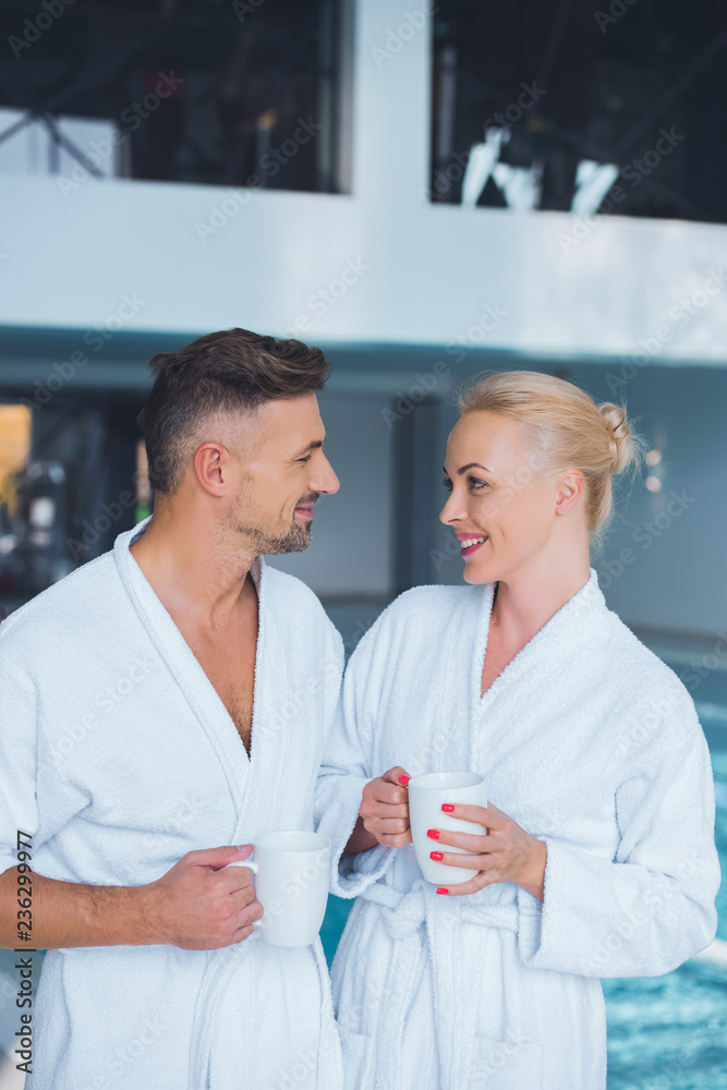 Obraz premium smiling couple in bathrobes standing near swimming pool with cups