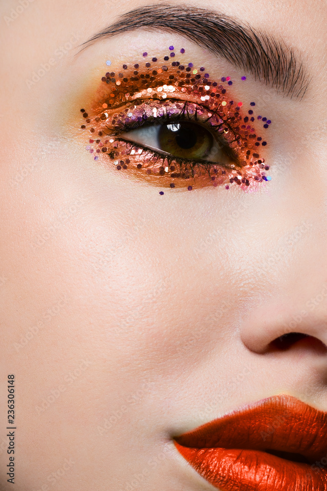 Fototapeta premium Close-up of the beauty of half a woman's face with creative fashionable make-up of sparkles. Black eyeliner and long eyelashes decorate hazel eyes and lips to match. Well-groomed skin after spa