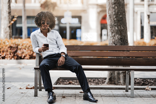 A curly young Asian businessman in sunglasses is sitting on the street bench, looking at the camera and holding a smartphone in his hand, with a copy space place for your logo, text message or advert