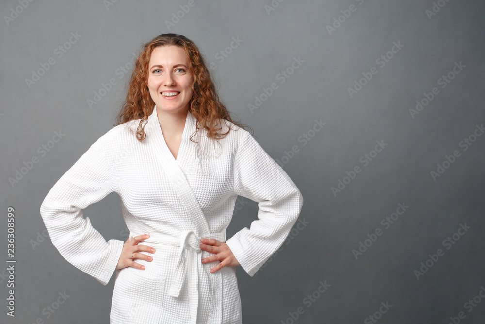Beautiful girl wearing white bathrobe, looking at camera and smiling, a model with light nude make-up, gray studio background, beauty photo, copy space Beautiful girl wearing white bathrobe, looking at camera and smiling, a model with light nude make-up, gray studio background, beauty photo, copy space
