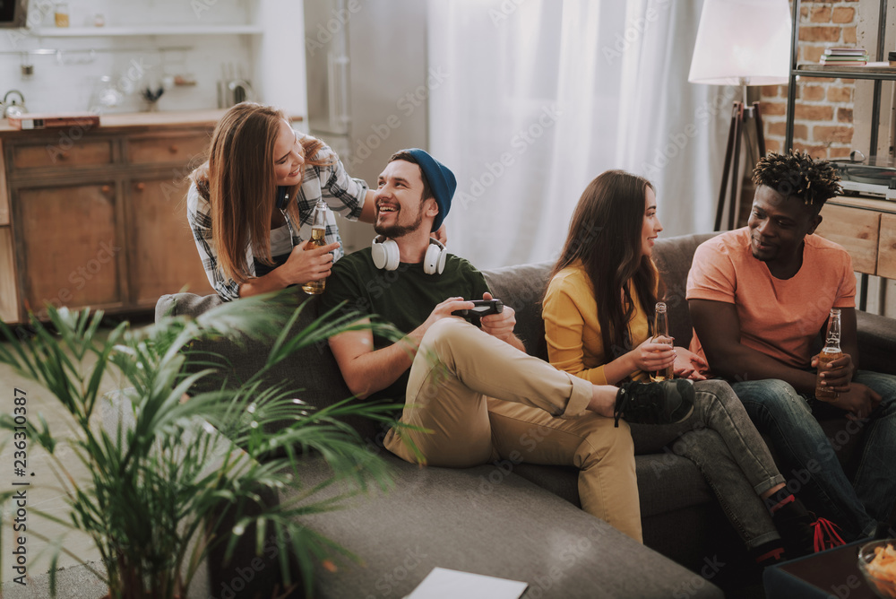 Naklejka premium Handsome bearded man in hat using joystick while charming lady looking at him and smiling. Joyful afro american guy sitting on couch and talking with brunette lady