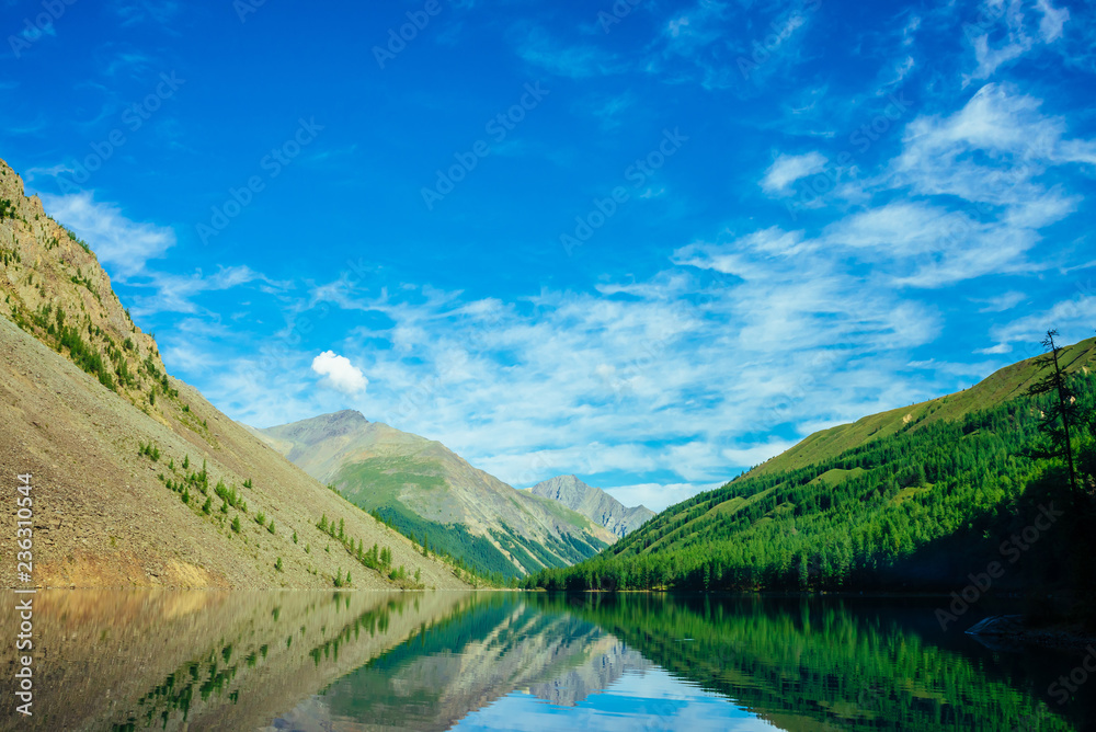 Wonderful mountain lake in valley of highlands. Giant mountains reflected in smooth clean water surface. Amazing coniferous forest in sunlight. Atmospheric vivid green landscape of majestic nature.