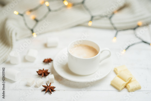 Coffee with milk, latte with cinnamon sticks and anise stars with white chocolate and marshmallow, on a light white background, with lights from a garland.